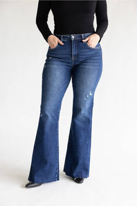 A person wearing dark blue high-waisted flare jeans with distressed details on the thighs, paired with a black long-sleeve top and black shoes, standing against a plain white studio background.