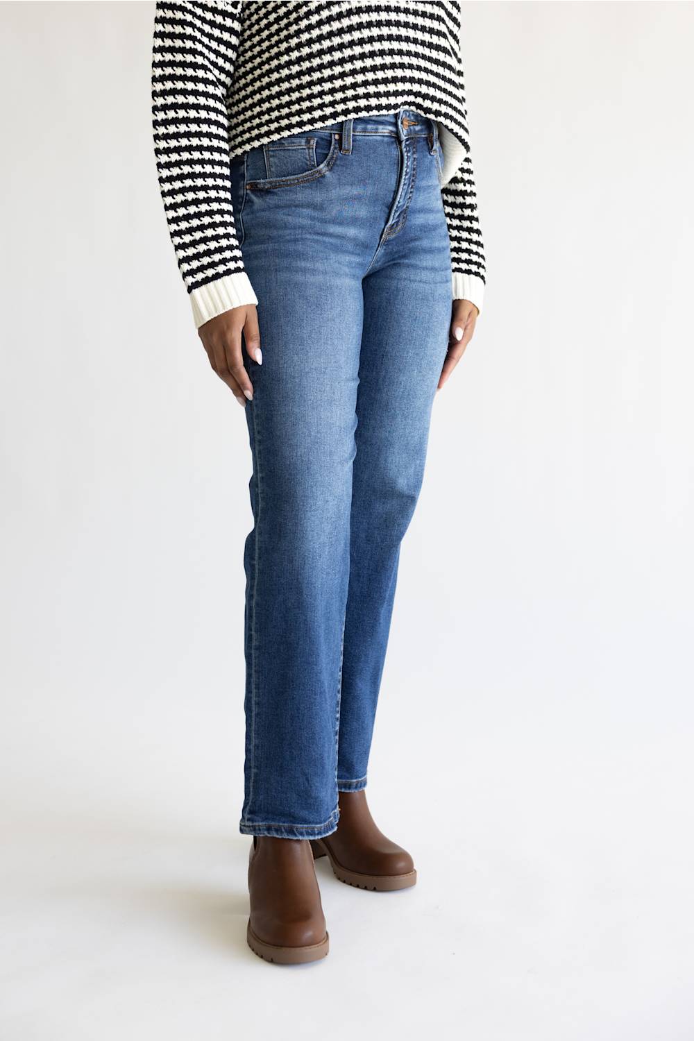 A person wearing medium-wash blue straight-leg jeans, a black and white houndstooth sweater, and brown ankle boots, standing against a plain white studio background.