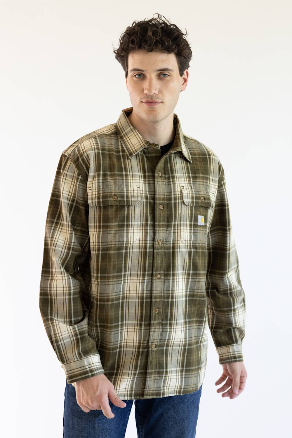 A man wearing a Carhartt long-sleeve plaid button-up shirt in olive green, beige, and brown tones, paired with blue jeans, photographed against a plain white studio background.