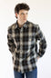 A man wearing a long-sleeve plaid button-up shirt by Carhartt, featuring a dark gray, black, and beige pattern with thin yellow lines, paired with blue jeans against a plain white background.