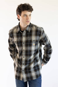 A man wearing a long-sleeve plaid button-up shirt by Carhartt, featuring a dark gray, black, and beige pattern with thin yellow lines, paired with blue jeans against a plain white background.