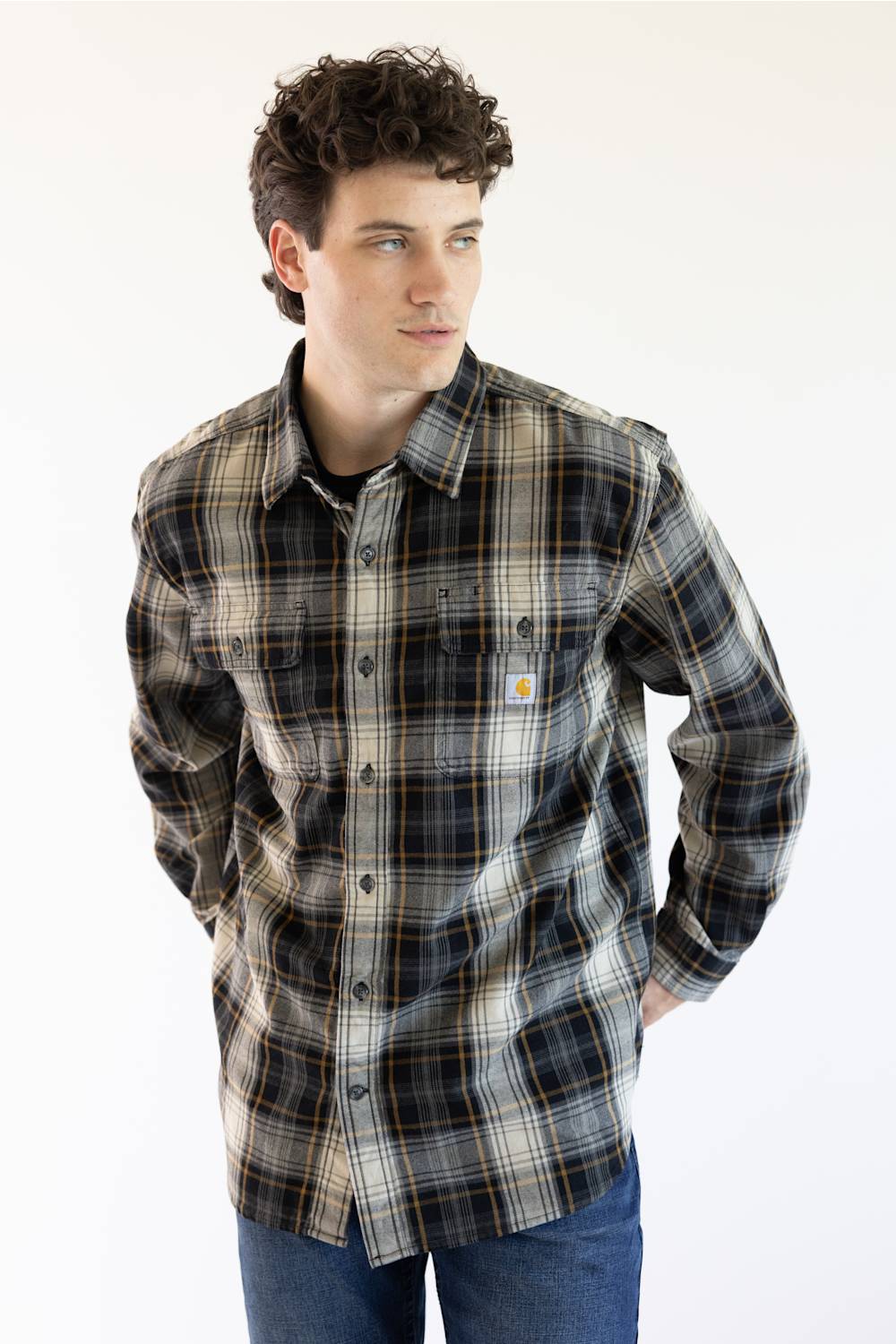 A man wearing a long-sleeve plaid button-up shirt by Carhartt, featuring a dark gray, black, and beige pattern with thin yellow lines, paired with blue jeans against a plain white background.