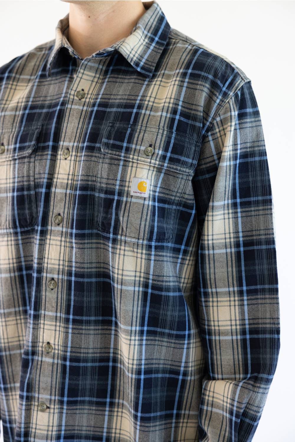 A close-up of a person wearing a Carhartt plaid button-up shirt. The shirt features a beige and navy blue plaid pattern with thin blue and white lines, and the signature yellow Carhartt logo is visible on the chest pocket.