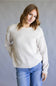 A woman wearing a light beige knit crewneck sweater with delicate ruffle trim at the collar and cuffs, paired with blue jeans, standing against a neutral gray studio background.