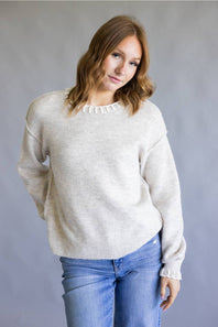 A woman wearing a light beige knit crewneck sweater with delicate ruffle trim at the collar and cuffs, paired with blue jeans, standing against a neutral gray studio background.
