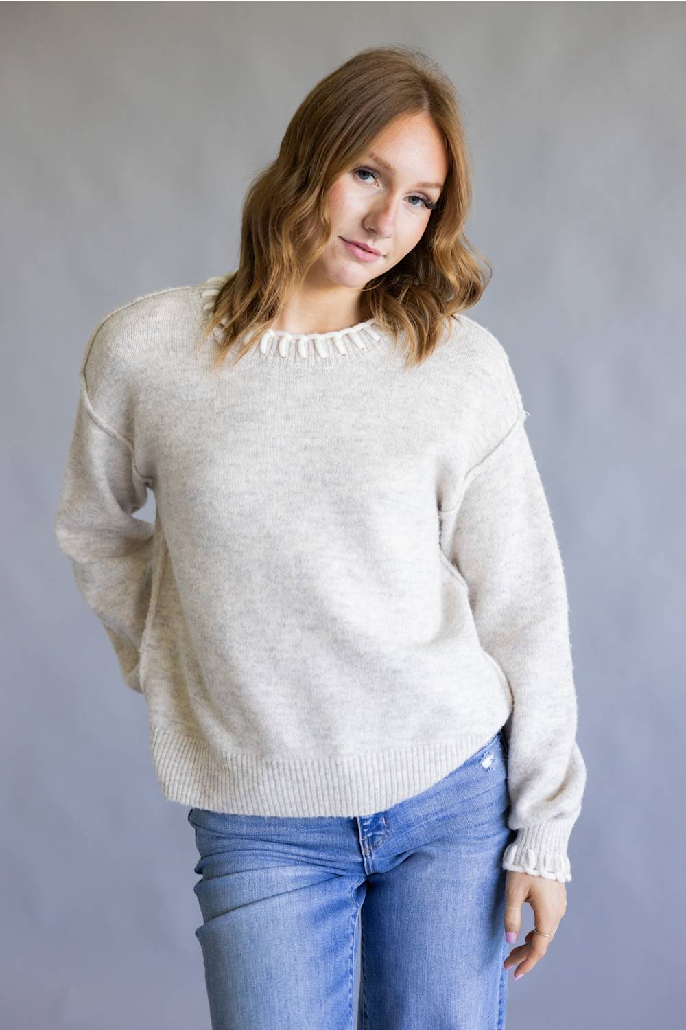 A woman wearing a light beige knit crewneck sweater with delicate ruffle trim at the collar and cuffs, paired with blue jeans, standing against a neutral gray studio background.