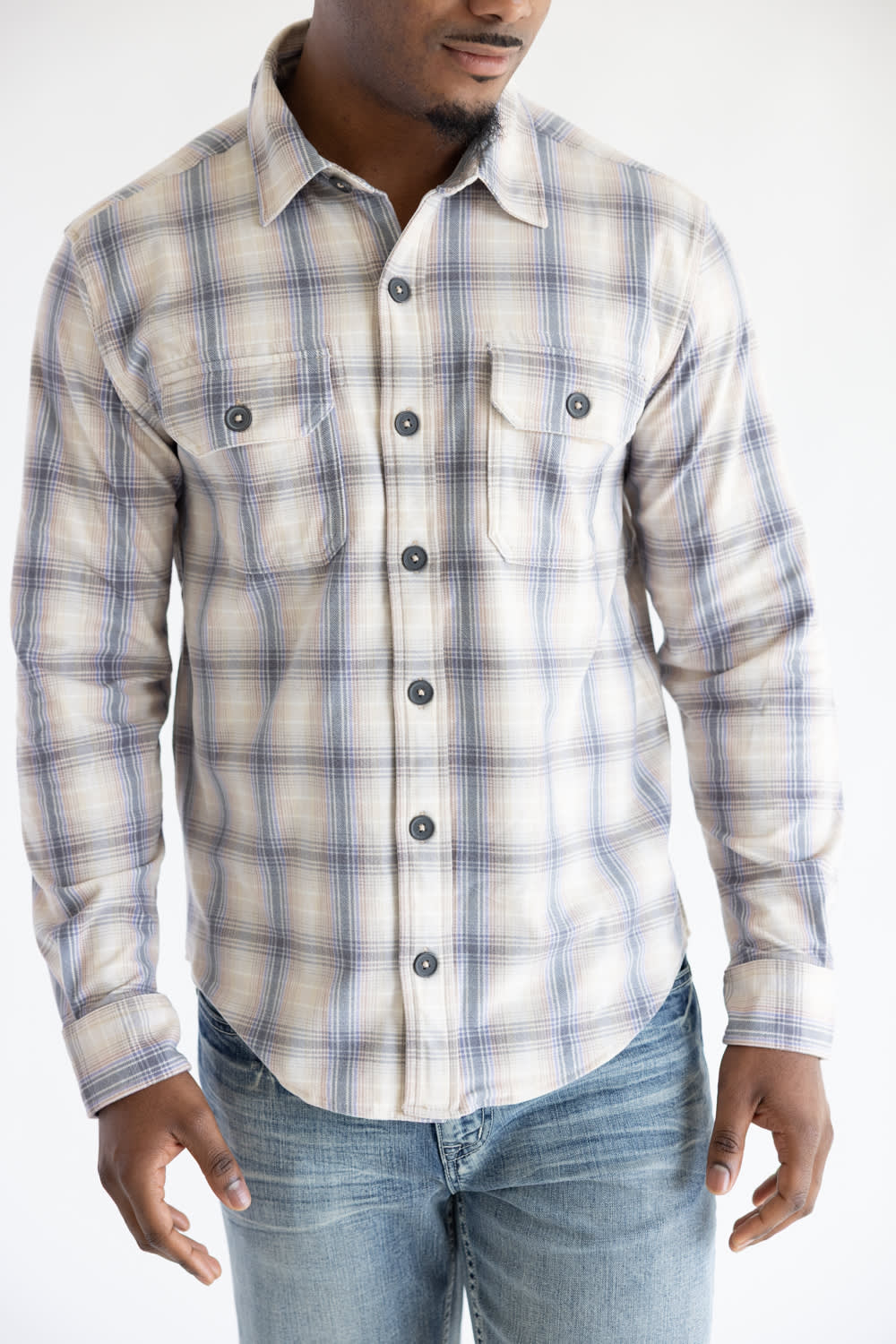 Cropped front-facing view of a man wearing the Hallstatt Knit Button Up Flannel Shirt in Sand with a blue, beige, and gray plaid pattern, paired with light-wash jeans.