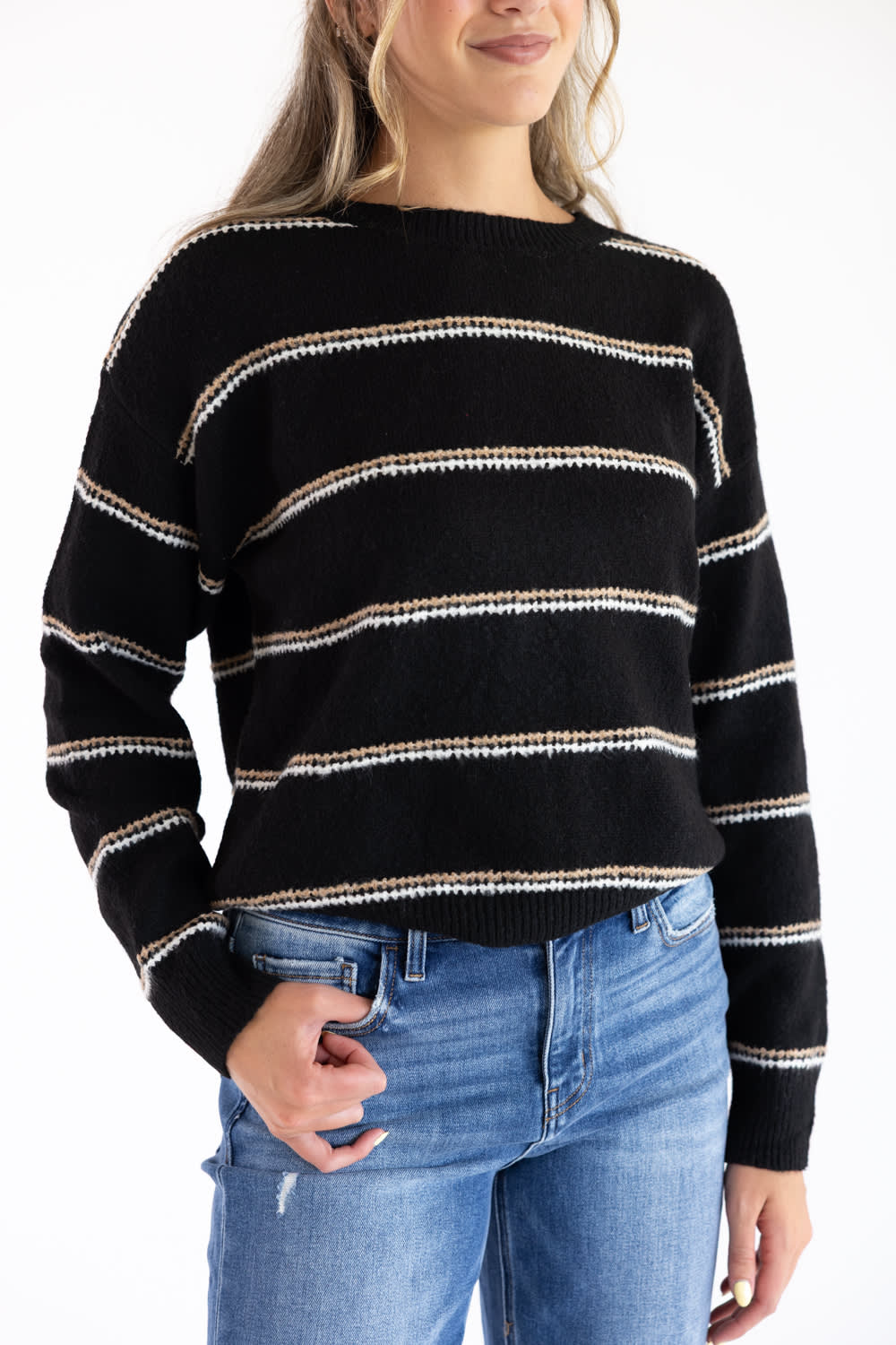 Model wearing a black crew neck knit sweater with thin tan and white embroidered horizontal stripes, ribbed neckline, and long sleeves paired with blue denim jeans.
