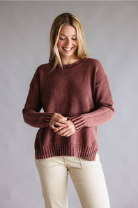 A woman wearing a dusty rose ribbed knit sweater and cream pants, standing against a neutral gray studio background.