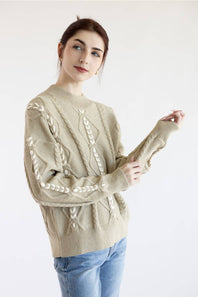 A woman wearing a beige cable-knit sweater with white decorative patterns and light blue jeans, posing against a plain white background.