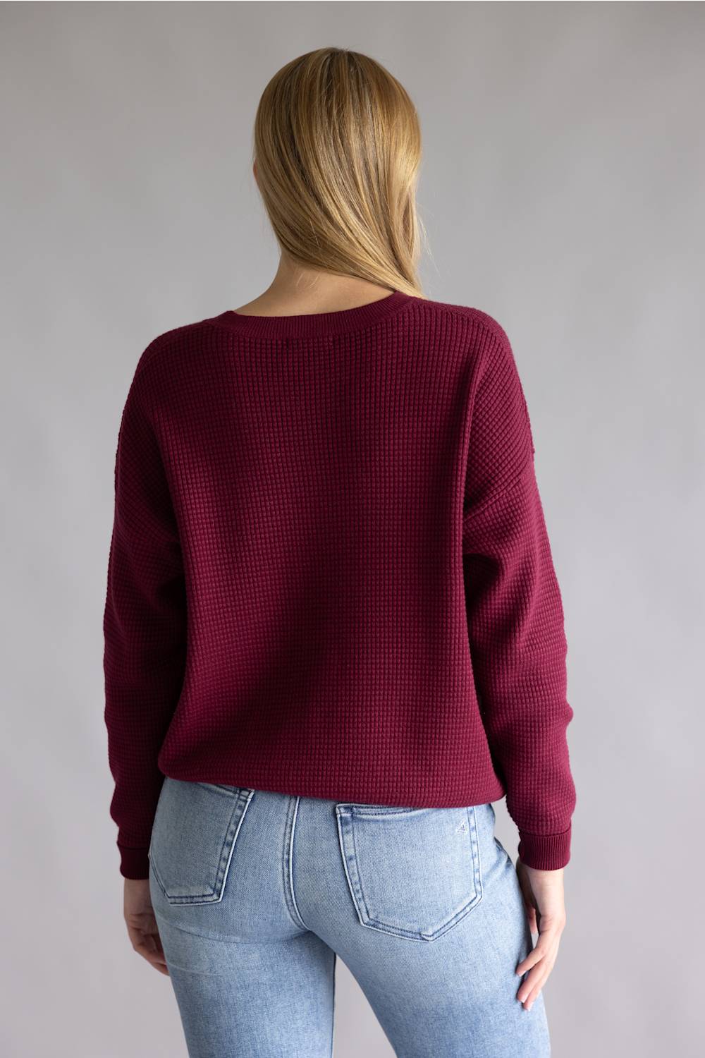 A woman viewed from behind wearing a textured, long-sleeve crewneck sweater in deep burgundy and light wash blue jeans, set against a plain gray studio background.