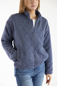 A woman wearing a heathered slate blue quilted zip-up jacket over a white top and blue jeans, shown from the waist up against a plain white background.