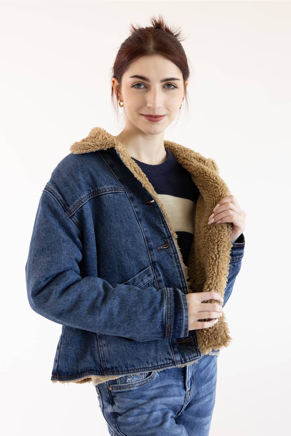 A woman wearing a blue denim jacket with a brown sherpa fleece lining, paired with a striped shirt and jeans, photographed against a plain white studio background.