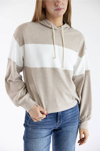 A woman wearing a beige and white color-blocked knit hoodie with drawstrings, paired with blue jeans, against a plain white background.