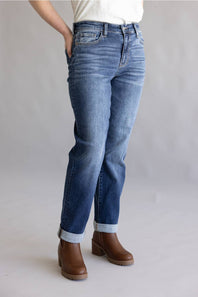A person wearing medium-wash blue denim jeans with rolled cuffs and brown ankle boots, shown from the waist down against a light gray studio background.