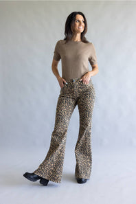 A woman modeling leopard-print flared pants and a beige short-sleeve top, standing against a neutral light gray studio backdrop.