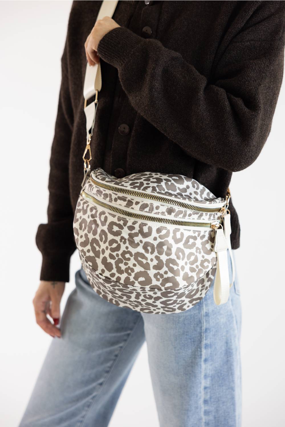 A person wearing a dark brown cardigan and light blue jeans carries a leopard-print crossbody bag with a beige strap and gold-tone hardware against a plain white background.