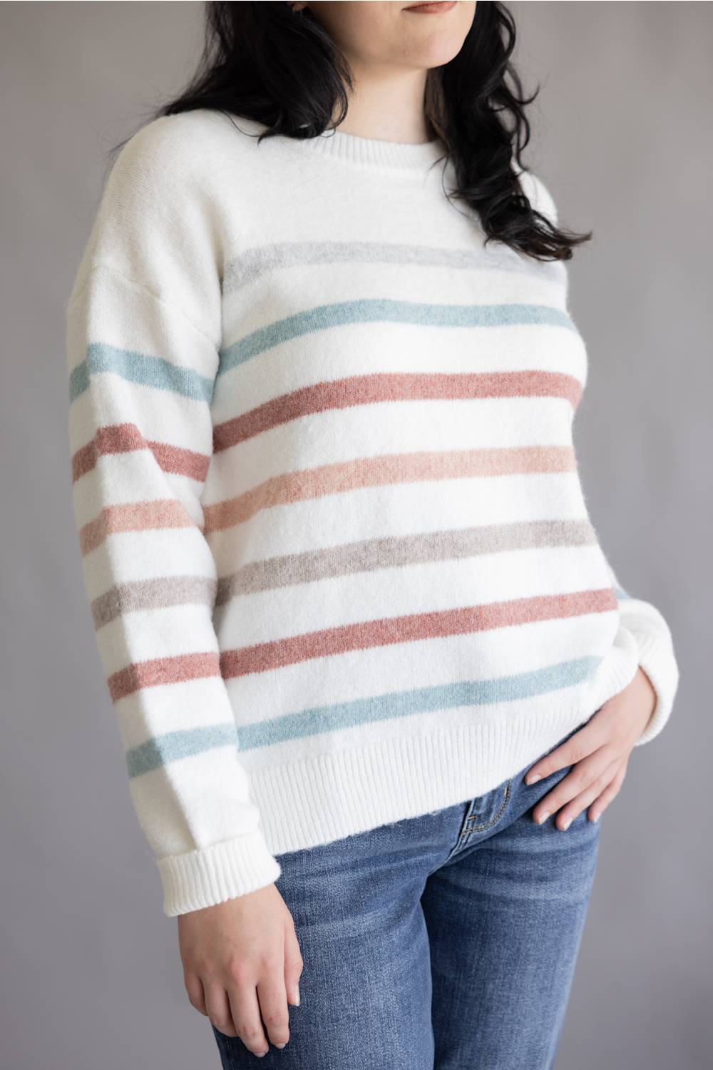 A woman wearing a cream knit sweater with horizontal stripes in dusty pink, light blue, and light gray, paired with blue jeans, shown from the neck down against a neutral gray background.