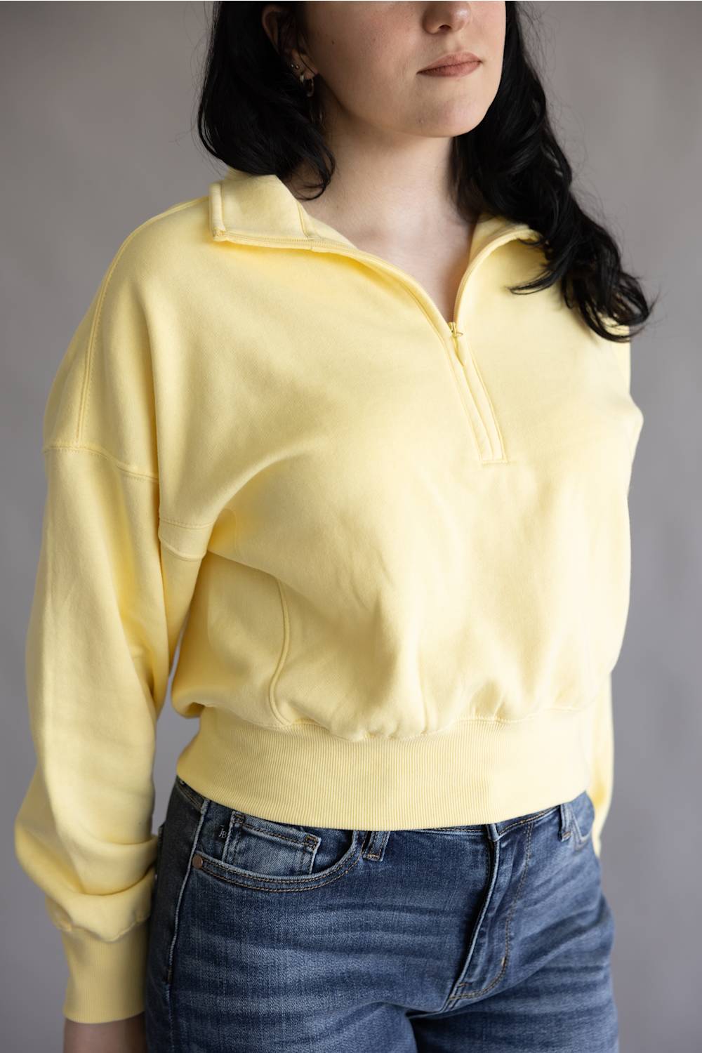 A woman wearing a pale yellow half-zip sweatshirt with a stand collar and blue jeans, shown from the chest down against a plain gray background.