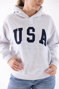 Close-up of a blonde woman wearing an ice grey Reflex fleece pullover hoodie with dark navy "USA" block lettering across the chest and a front kangaroo pocket.