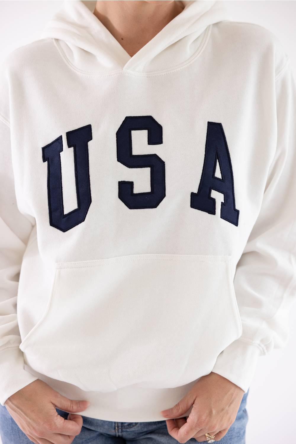 Close-up of an ivory Reflex fleece pullover hoodie featuring dark navy blue "USA" block letters across the chest and a front kangaroo pocket.