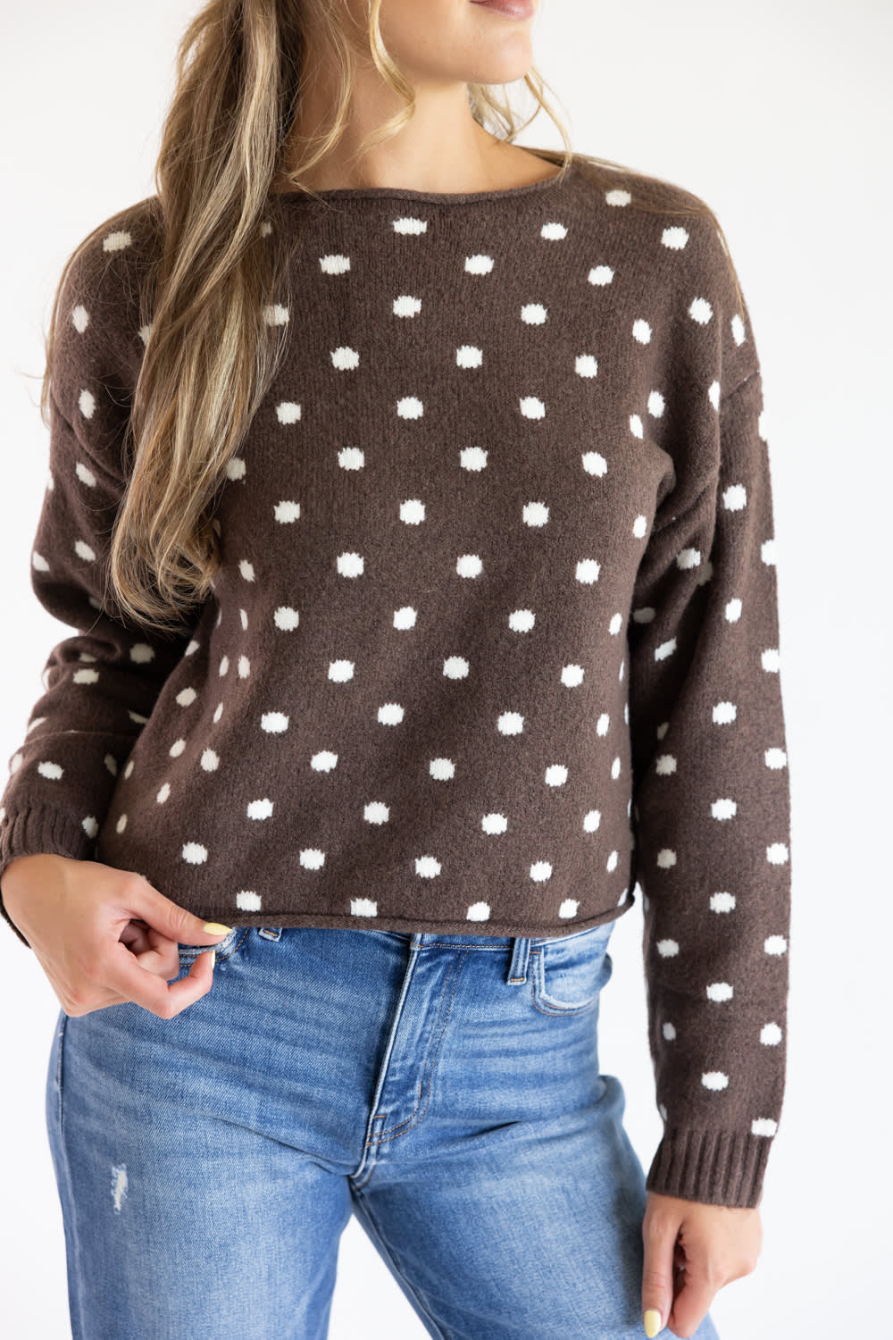 Close-up front view of a model wearing the Just Polly dark brown long-sleeve knit sweater featuring an ivory polka dot pattern, a slightly rolled boat neckline, and ribbed cuffs against a plain light background.