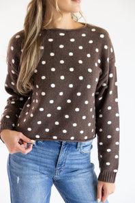 Close-up front view of a model wearing the Just Polly dark brown long-sleeve knit sweater featuring an ivory polka dot pattern, a slightly rolled boat neckline, and ribbed cuffs against a plain light background.