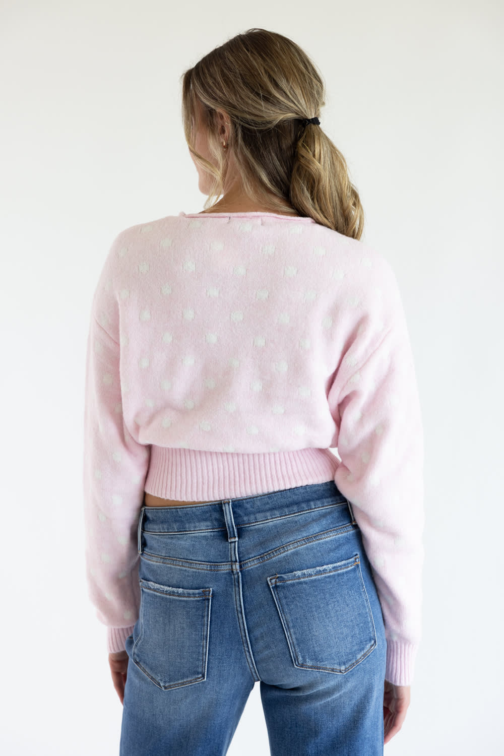 Back view of a model wearing the Just Polly light pink long-sleeve knit sweater featuring a white polka dot pattern, a slightly rolled boat neckline, and a wide ribbed waistband, styled with denim jeans against a plain white background.