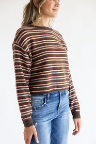 Side-profile view of a model wearing the Just Polly long-sleeve knit sweater featuring horizontal stripes in brown, red, and tan, a slightly rolled boat neckline, and ribbed cuffs, styled with denim jeans against a plain white background.