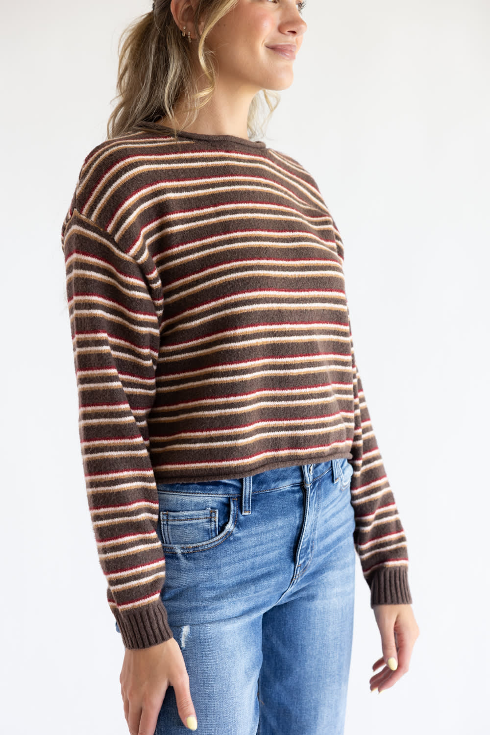 Side-profile view of a model wearing the Just Polly long-sleeve knit sweater featuring horizontal stripes in brown, red, and tan, a slightly rolled boat neckline, and ribbed cuffs, styled with denim jeans against a plain white background.