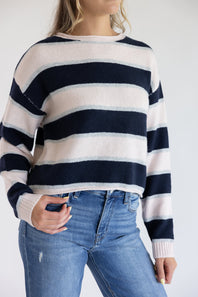 Close-up front view of a model wearing the Just Polly long-sleeve knit sweater featuring wide light pink and navy horizontal stripes with thin heather grey accents, a slightly rolled boat neckline, and ribbed cuffs against a plain background.
