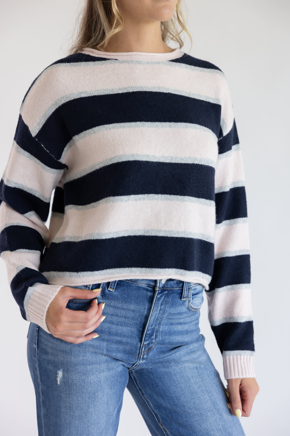 Close-up front view of a model wearing the Just Polly long-sleeve knit sweater featuring wide light pink and navy horizontal stripes with thin heather grey accents, a slightly rolled boat neckline, and ribbed cuffs against a plain background.