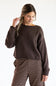 A model wearing the Just Polly Boat Neck Sweater in dark brown, paired with brown and white striped trousers against a plain off-white background.