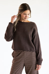 A model wearing the Just Polly Boat Neck Sweater in dark brown, paired with brown and white striped trousers against a plain off-white background.