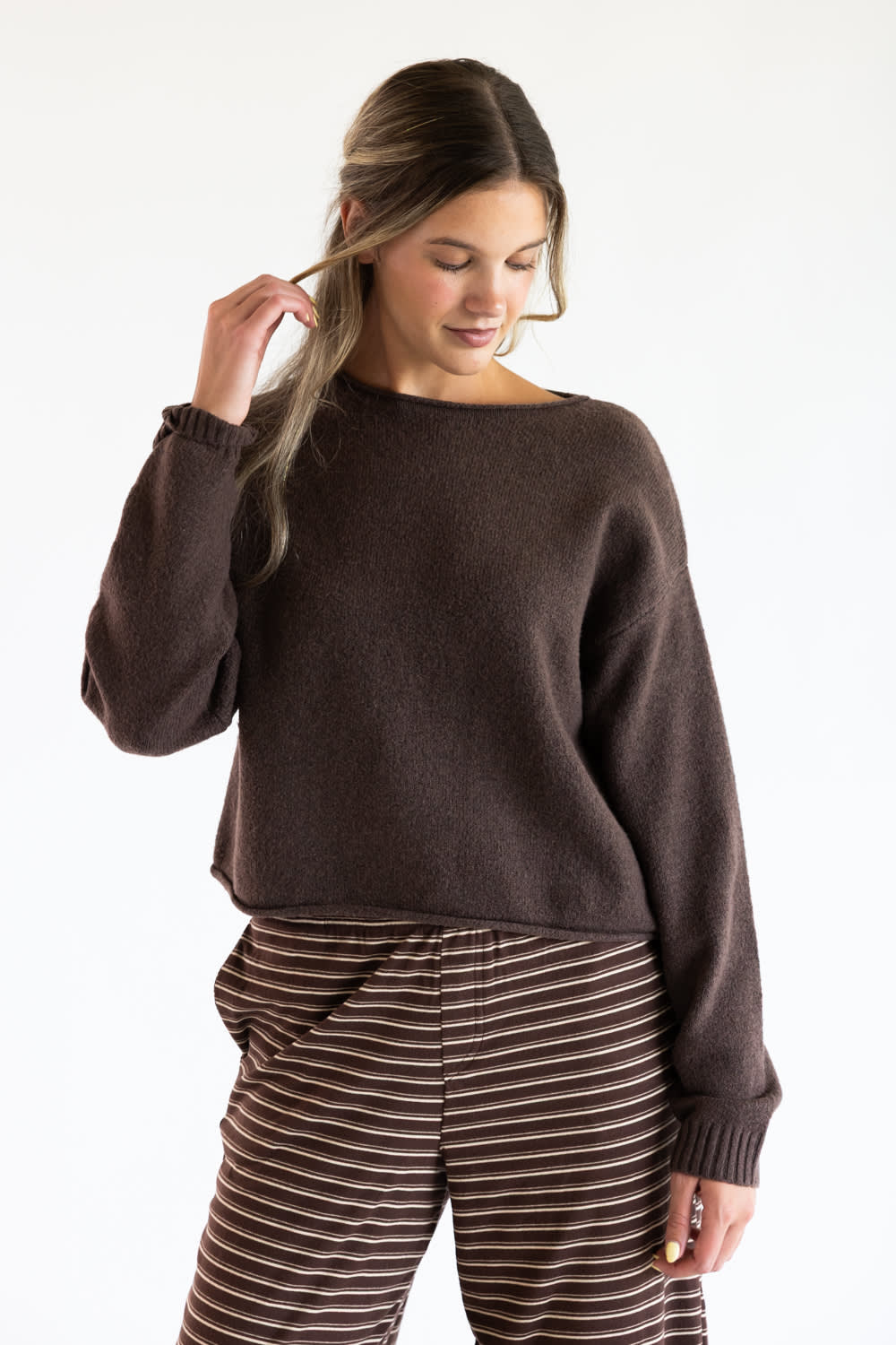 A model wearing the Just Polly Boat Neck Sweater in dark brown, paired with brown and white striped trousers against a plain off-white background.