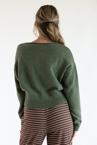 Back view of a model wearing the Just Polly olive green long-sleeve knit sweater featuring a wide ribbed waistband and cuffs, styled with brown and white striped pants against a plain white background.