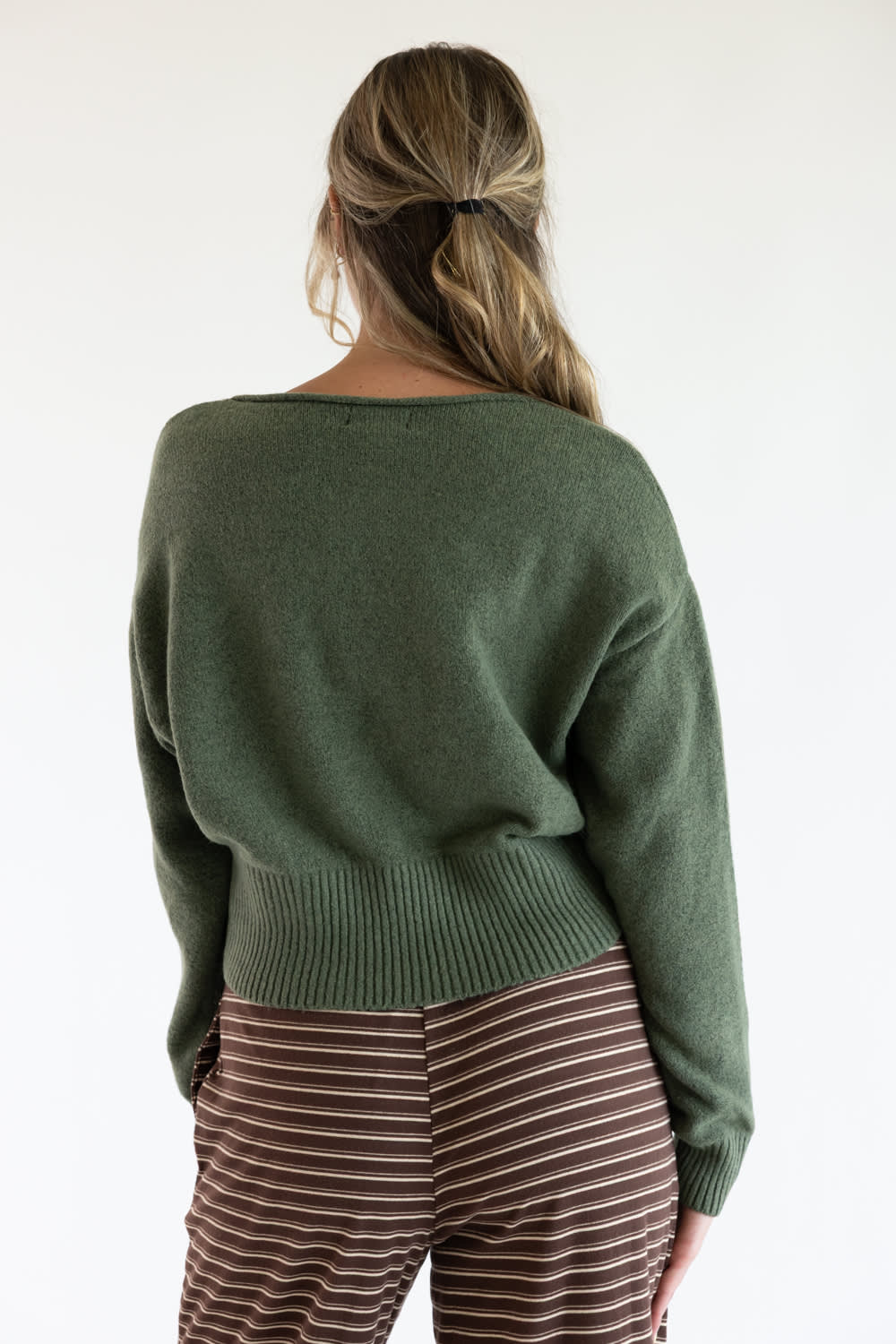 Back view of a model wearing the Just Polly olive green long-sleeve knit sweater featuring a wide ribbed waistband and cuffs, styled with brown and white striped pants against a plain white background.