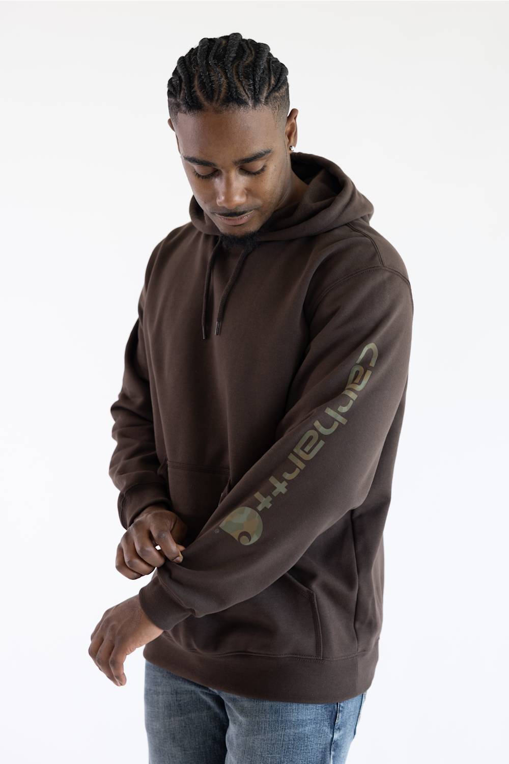 Three-quarter profile view of a male model against a white background, wearing a dark clove brown Carhartt hooded sweatshirt with "carhartt" printed down the left sleeve, paired with medium wash denim jeans.