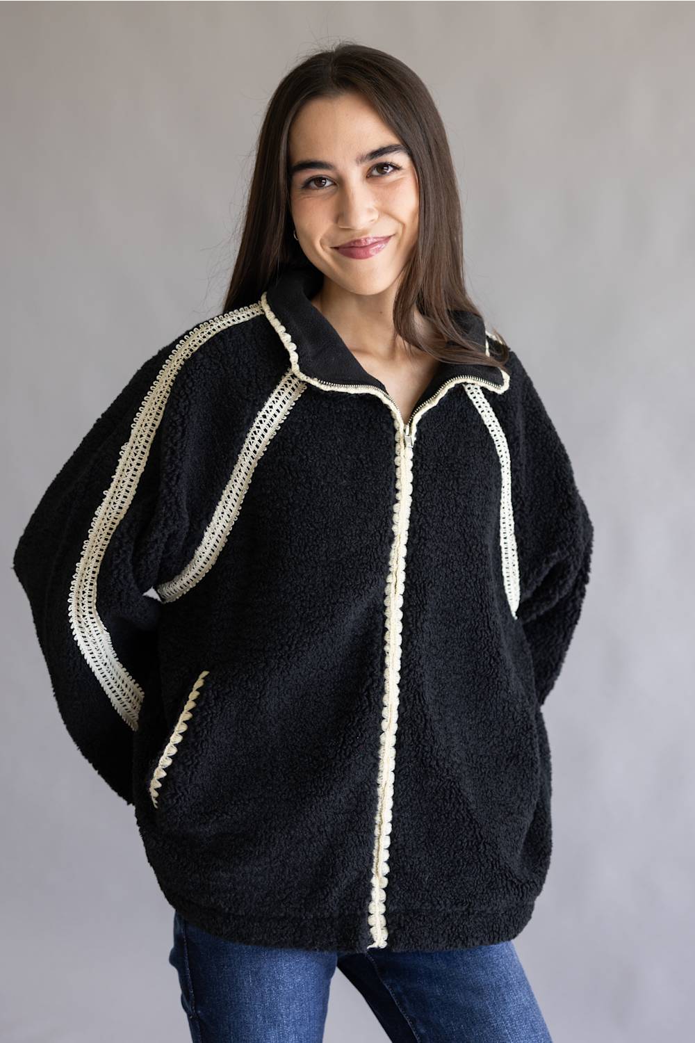 A woman wearing a black fuzzy zip-up jacket with cream-colored trim along the zipper, collar, and sleeve seams, paired with blue jeans, set against a plain gray studio background.