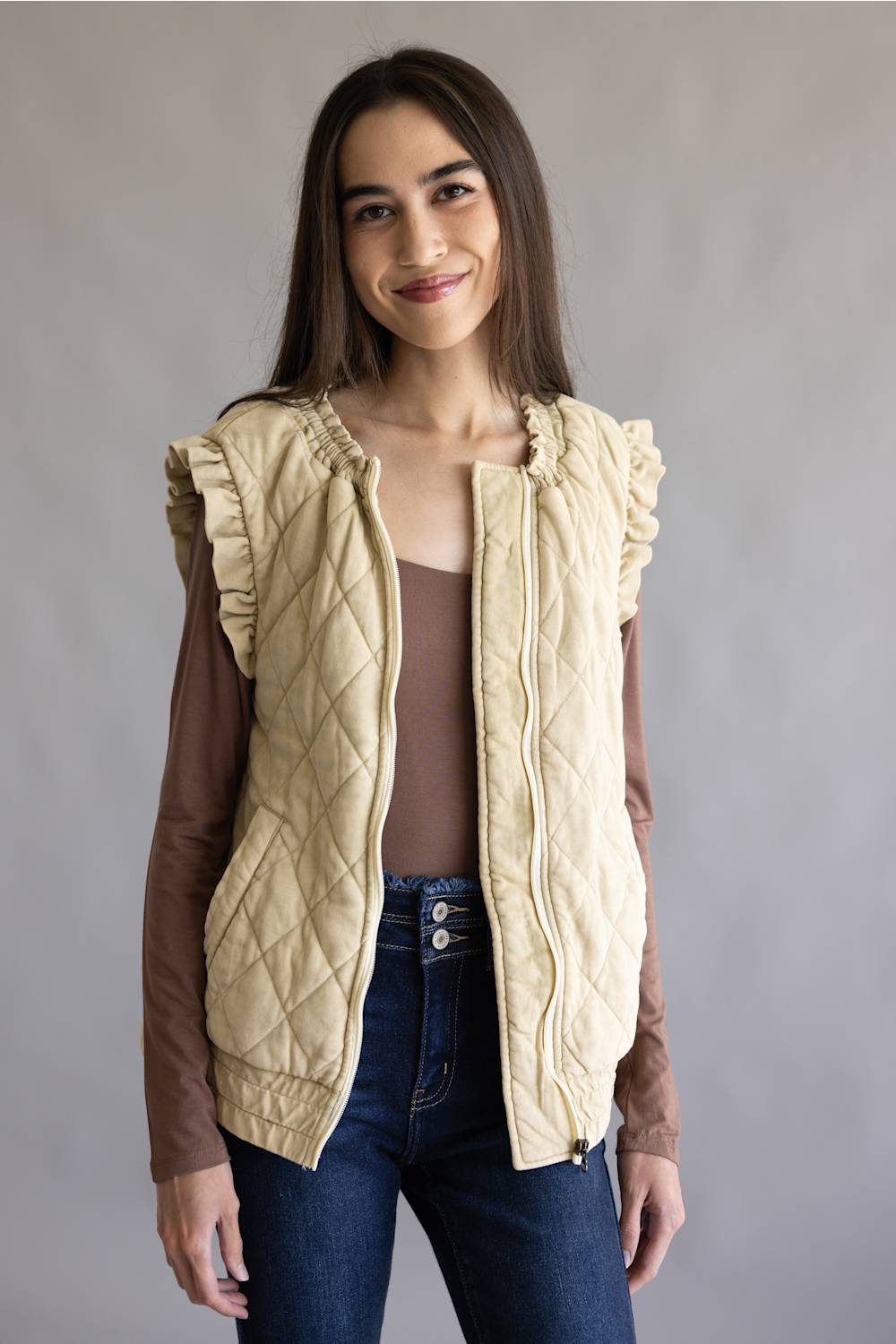 A woman modeling a sleeveless quilted vest in a warm cream-beige color, styled over a brown long-sleeve top and blue jeans against a plain gray studio background.