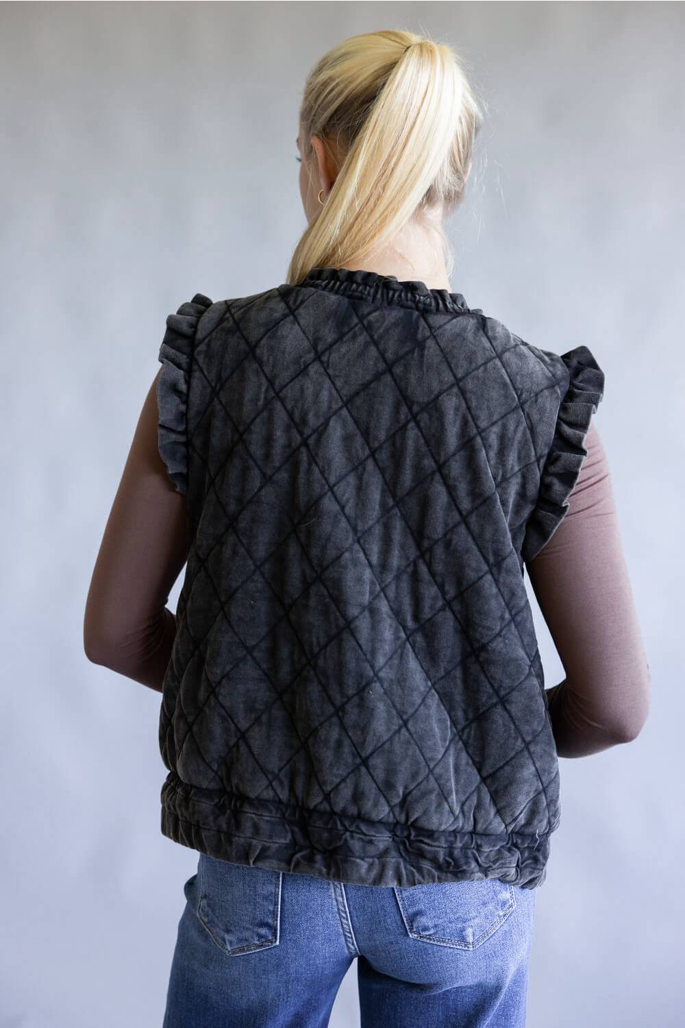 A woman viewed from behind wearing a sleeveless black quilted vest with ruffled trim over a mauve-brown top and blue jeans, against a plain light gray background.