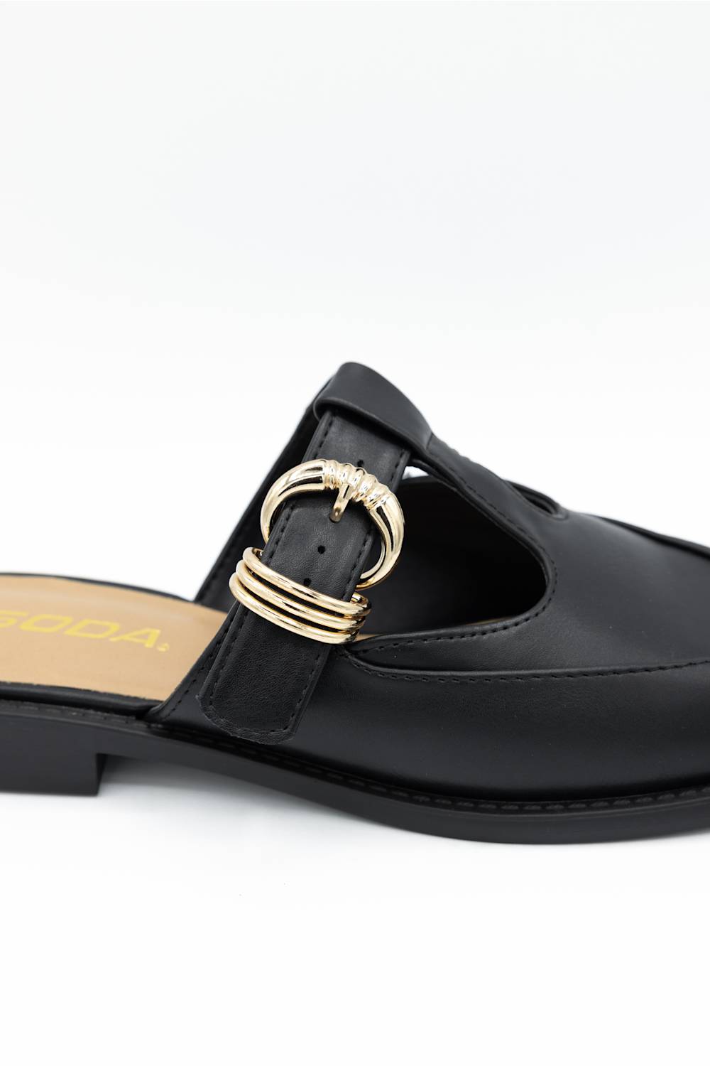 A close-up of a black leather mule sandal featuring a decorative gold-tone metal ring buckle on the strap, set against a plain white background.