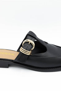 A close-up of a black leather mule sandal featuring a decorative gold-tone metal ring buckle on the strap, set against a plain white background.