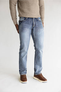 A person wearing light-wash blue jeans with a beige knit sweater and brown shoes, shown from the chest down against a plain light gray studio background.