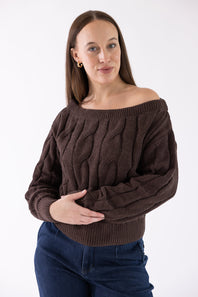 A woman wearing a dark brown off-the-shoulder cable-knit sweater and dark blue jeans, standing against a plain white studio background.