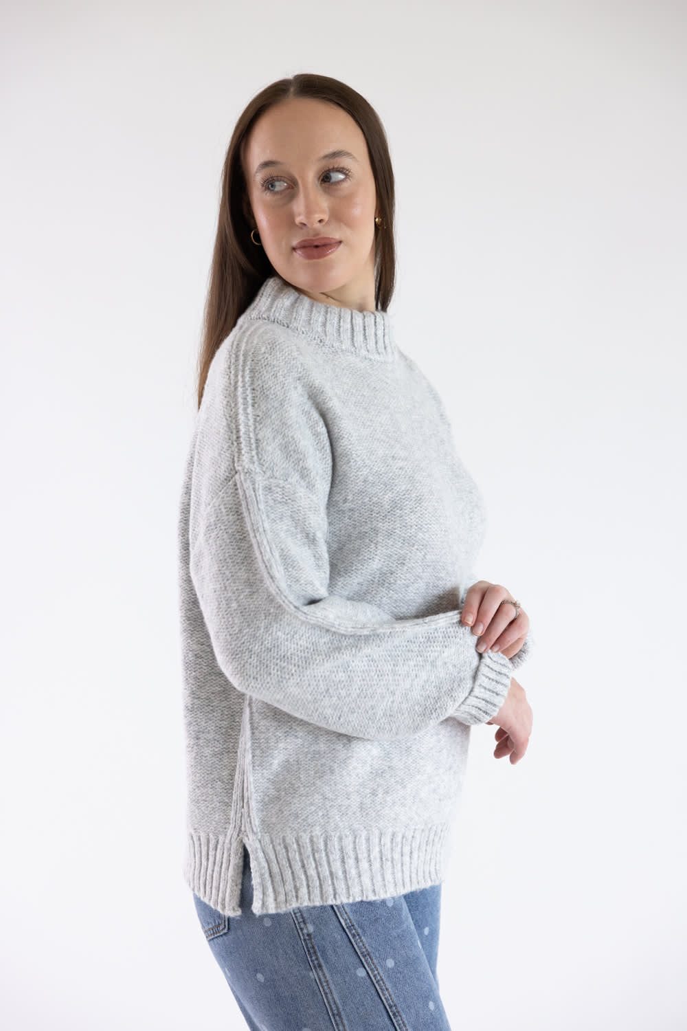 A woman wearing a light gray knit sweater and blue jeans, standing against a plain white studio background.