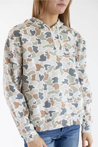 A woman wearing a camouflage hoodie with a beige, brown, gray, and green pattern, paired with blue jeans, shown against a plain white background.