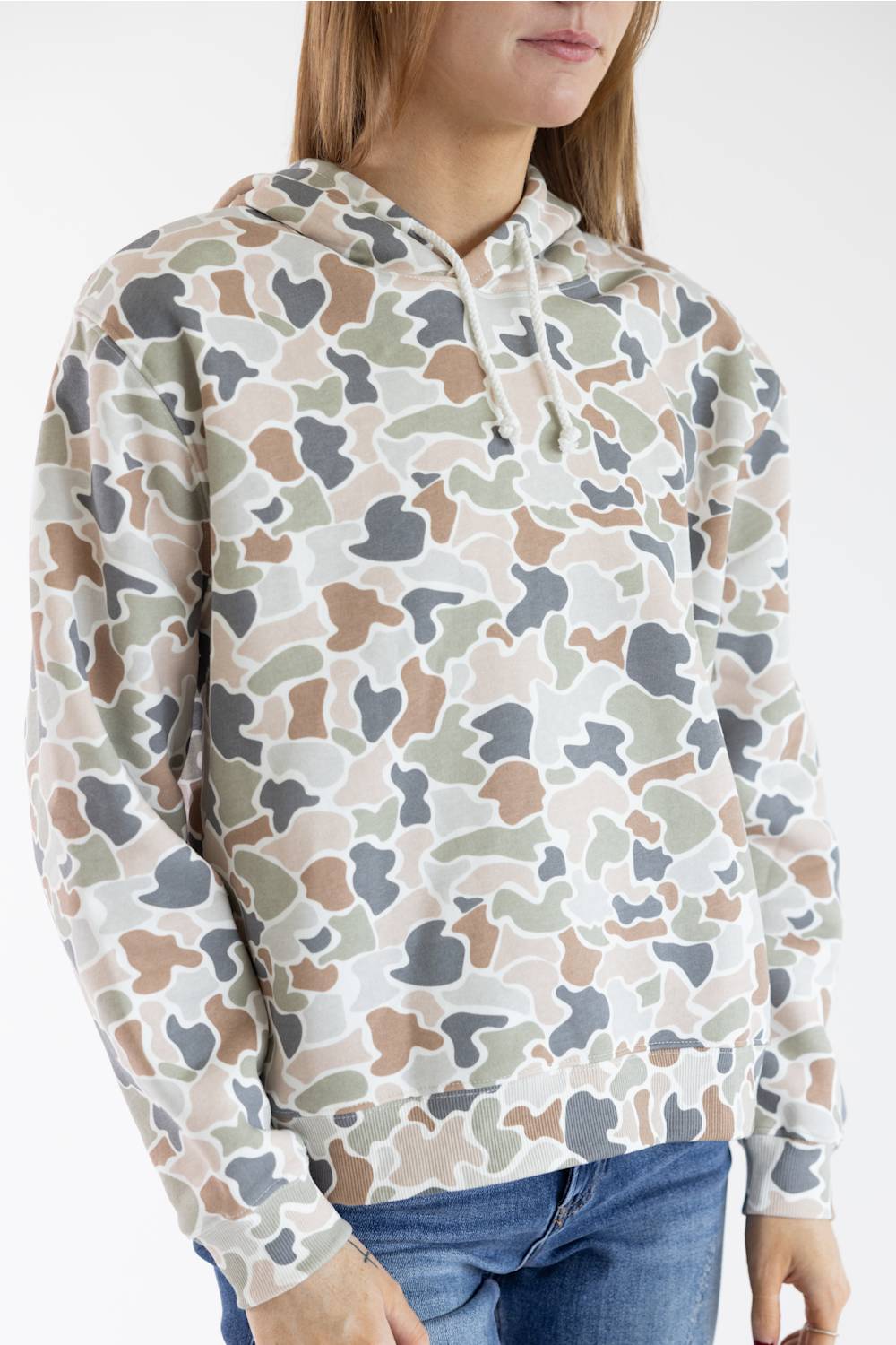 A woman wearing a camouflage hoodie with a beige, brown, gray, and green pattern, paired with blue jeans, shown against a plain white background.