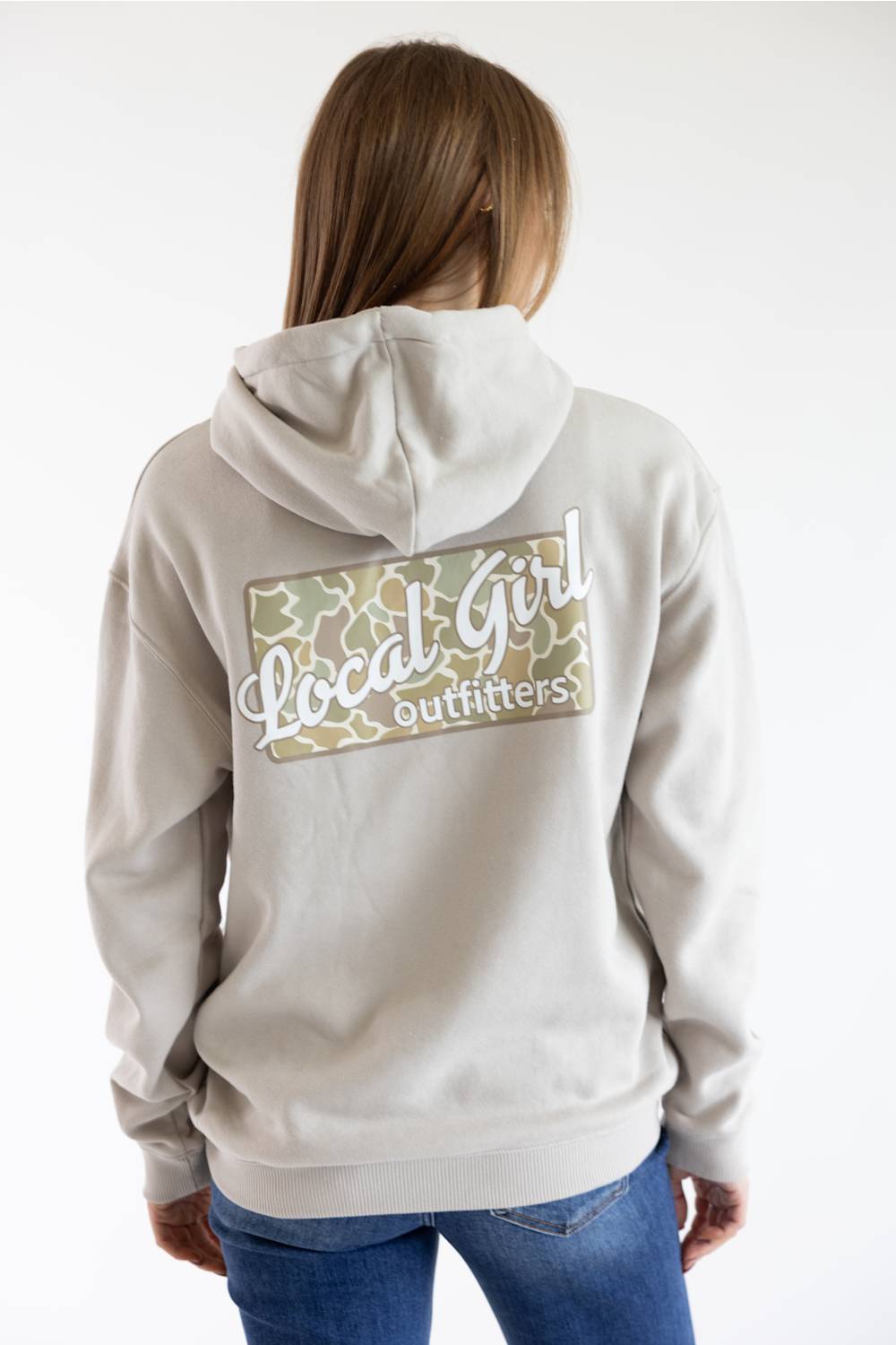 A woman viewed from behind wearing a light beige hoodie with a large back graphic that reads 'Local Girl Outfitters' over a green and beige patterned rectangle, paired with blue jeans against a plain white background.