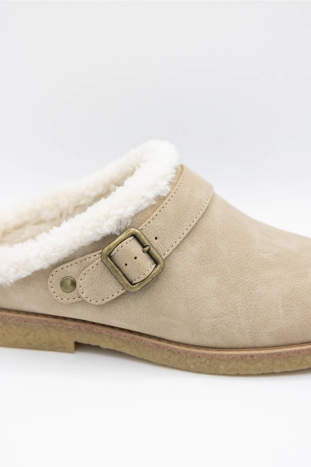 A close-up of a beige suede clog-style slipper with a white sherpa lining and an antique bronze buckle strap, shown against a plain white background.